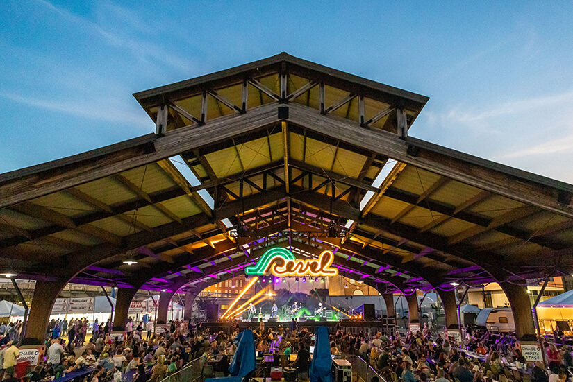 Wide photo of the pavilion at Festival Plaza in Shreveport, Louisiana. Under the pavilion are hundreds of people watching a live band. Behind the band is a large neon sign with the word "Revel" on it.
